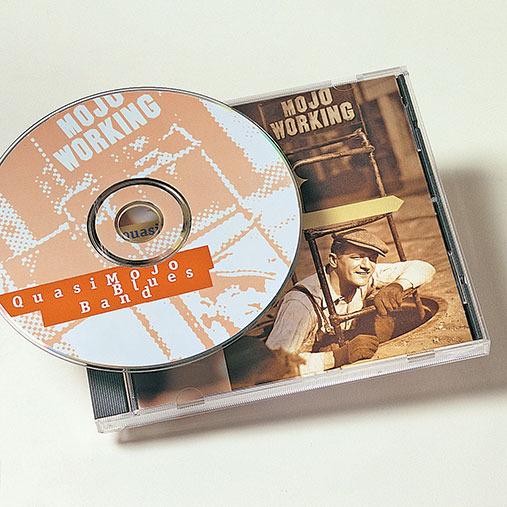 CD and jewel case for the album “Mojo Working” by the Quasi Mojo Blues Band. The CD features a stylized orange and white graphic design, while the cover art shows a sepia-toned photo of a smiling man in vintage workwear kneeling on the ground near an industrial drum. The design blends retro and blues themes.