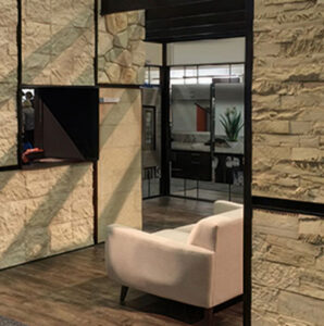 A Creative Mines trade show booth designed with large stone veneer panels in varying textures and tones. The structure includes black-framed divisions and a wood-style floor, with a cream-colored modern lounge chair inside. A small monitor is embedded in one of the walls. At the top, illuminated initials “CM” are mounted above a metal mesh structure, serving as a branded identifier visible from afar. The surrounding booths and signage hint at a bustling convention environment.