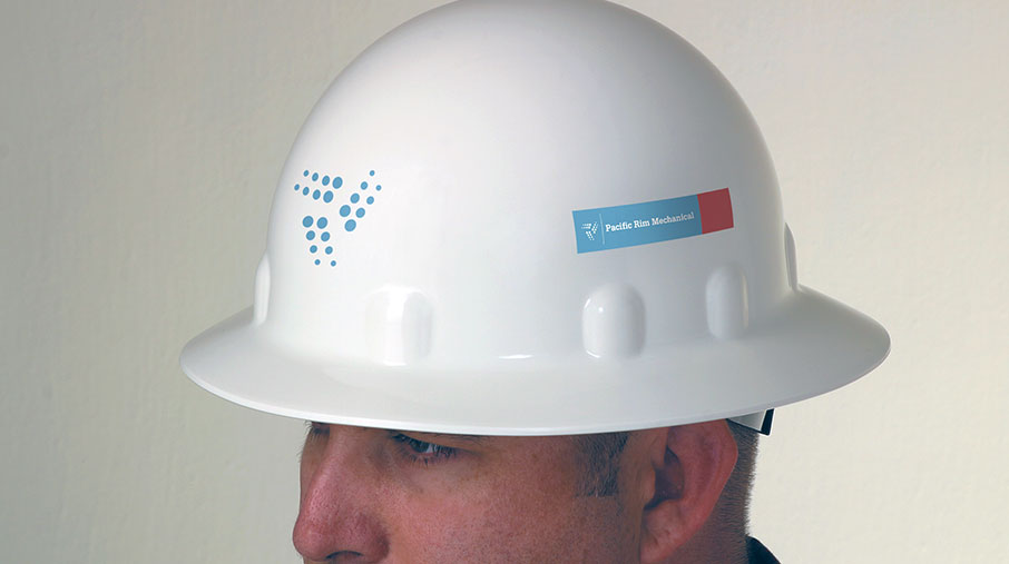 A close-up view of a white hard hat worn by a worker, featuring PacRim Mechanical branding. The front of the helmet displays the company’s dotted triangle logo in blue, while the side features a rectangular label with the full company name “Pacific Rim Mechanical” on a blue background with an orange accent. The hard hat signifies safety and professionalism in an industrial or construction environment.