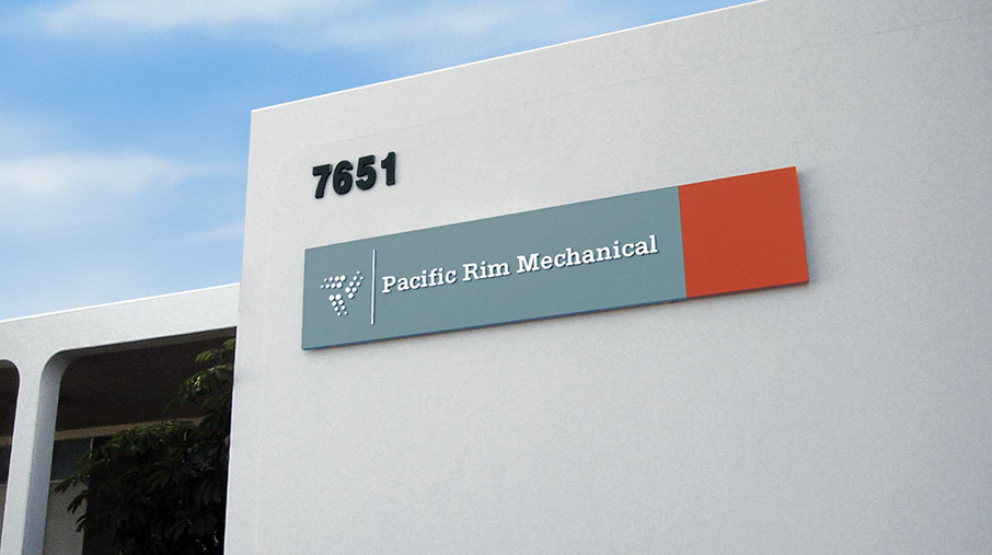 Exterior signage for Pacific Rim Mechanical mounted on a clean white building facade. The rectangular sign features a blue-gray background with the company name “Pacific Rim Mechanical” in white serif text, accompanied by the triangular PacRim logo made of small dots. An orange accent block adds contrast on the right side of the sign. The building number “7651” is displayed in black above the sign, and part of an overhang and tree can be seen on the left. The sky is bright and partly cloudy.