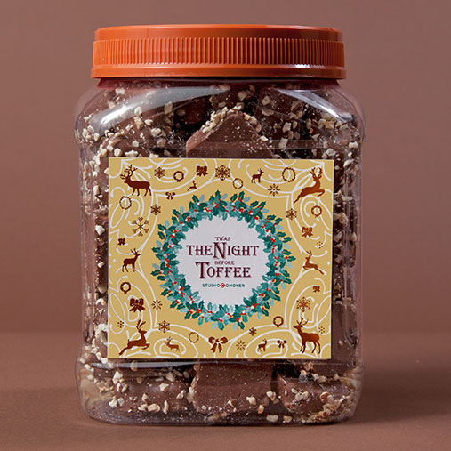 A clear plastic jar filled with chunks of chocolate-covered toffee, topped with an orange screw-on lid. The festive label on the front reads “The Night Before Toffee” by StudioConover and features a holiday design with reindeer, bows, and a wreath motif on a golden background.