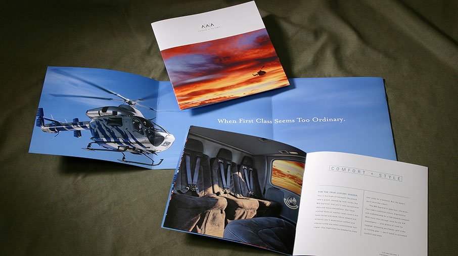 Printed marketing materials for Triple A featuring luxury helicopter services. The brochures are laid out on a green fabric surface and display vivid imagery of a helicopter in flight, a plush interior cabin with leather seating, and a dramatic sunset sky. Visible text includes the tagline “When First Class Seems Too Ordinary” and section heading “Comfort + Style.”