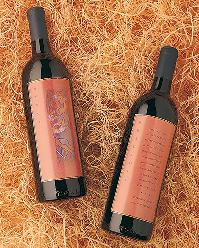 Two wine bottles with elegant copper-colored labels placed on a bed of natural raffia or straw. One label features a stylized abstract illustration of a face, while the other contains text describing the wine. The bottles have dark glass and black foil tops, suggesting a premium or boutique wine brand.