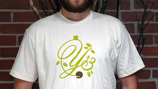 Person wearing a white T-shirt featuring the Y13 logo in light green script, styled with decorative swirls and leaf accents. The shirt is worn against a brick wall background.