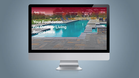 Computer monitor displaying the Angelus Paving Stones website homepage, featuring a backyard poolside setting with stylish concrete pavers and the headline text “Your Foundation For Outdoor Living Begins Here” overlaid on the scene.